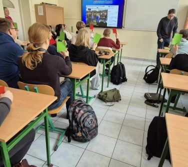 Sala szkolna, dzień. W ławkach siedzą nastolatkowie, podnoszą zielone i czerwone karteczki. Na przodzie ekran z wyświetlaną...