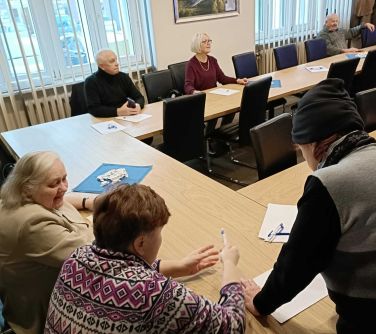 Sala konferencyjna, dzień. Na krzesłach przy stole w literkę U siedzą seniorzy