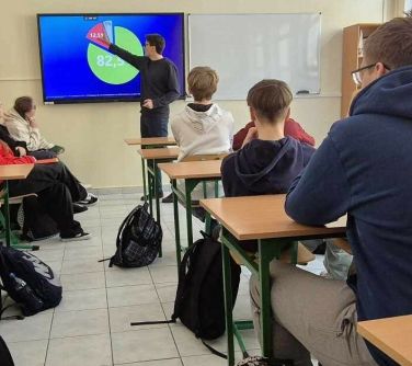 Oświetlona sala lekcyjna. Uczniowie w ławkach. Na przodzie na ekranie wyświetlany wykre. Mężczyzna wskazuje na dane