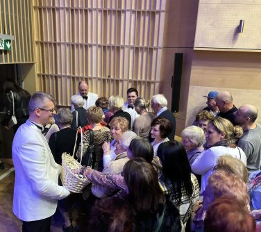 oświetlona sala koncertowa. Publicznośc opuszcza salę. Mężczyźni w białych marynarkach rozdają z koszyków upominki