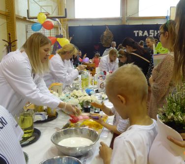 Jasna sala. Przy stołach osoby robią eksperymenty. Po lewej w białych fartuchach kobiety prowadzą eksperymenty