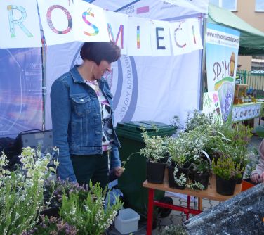 Na dworze, dzień. Przed budynkiem. Pod jasnym namiotem kobieta. Na przodzie na stolikach roślinki