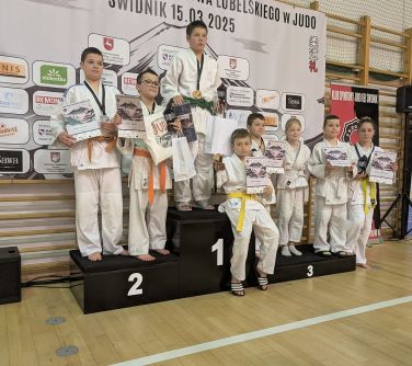 Hala sportowa. Na podium grupa zwycięzców w białych kimonach z dyplomami w dłoniach. W tle baner zawieszony na drewnianych...