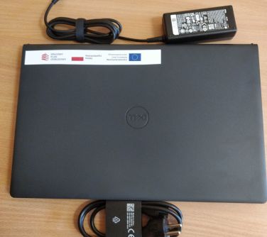 Na jasno-brązowym blacie leży złożony laptop z naklejką z logami. Na górze zasilacz. Poniżej złożony kabel
