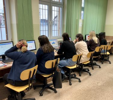 Oświetlona sala komputerowa. Sześć kobiet siedzi na krzeslach przed monitorami komputerowymi. Przed nimi okna, dzień
