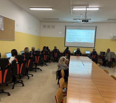 Oświetlona sala komputerowa. Na fotelach przed komputerami siedzą kobiety. Stoły ustawione w literę L. Naprzodzie ekran z...
