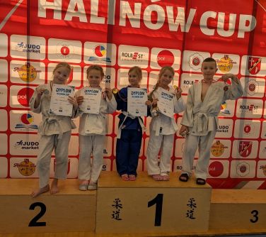 Oświetlona hala sportowa, dzień. Na drewnianym podium piątka dzieci w kimonach. Cztery dziewczynki trzymają dyplomy...