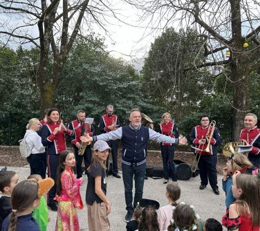 Na dworze, dzień. Po środku mężcyzzna z rozłożonymi na boki rękoma. Za nim w półkolu orkiestra dęta w mundrurach. W tle...
