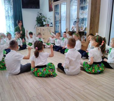 Jasne pomieszczenie. Na jasnej, drewnianej podłodze siedza dzieci elegancko wiosennie ubrane. Wśród nich kobieta ubrana na...
