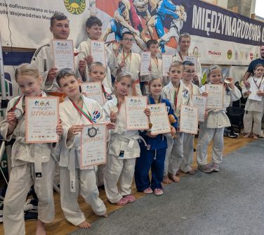 Jasna hala sportowa. Na tle banerów grupa judowków w białych strojach. W rękach trzymają dyplomy
