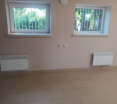 Jasne pomieszczenie. Beżowe ściany. Dwa okna z kratami. Za oknem dzień. Na dole na ścianach dwa mniejsze grzejniki