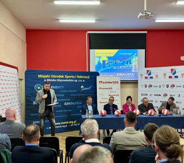 Oświetlona sala. Na przodzie na tle banerów przy stole siedzi pięć osób. Na stole biało-czerwone piłki do nogi. Po lewej do...