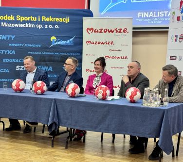 Oświetlona sala.Na tle banerów przy stole siedzi pięć osób. Na stole pięć biało-czerwonych piłek