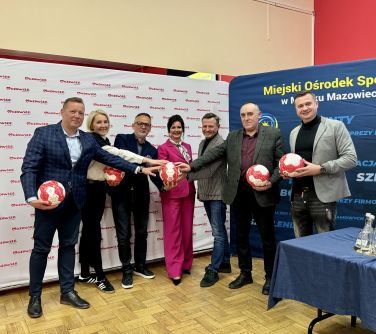 Oświetlona sala. Na tle banerów siedem osób trzyma piłki do nogi w dłoniach. Po środku jedna piłka do której każdy...