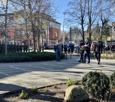 Na zewnątrz, dzień. Po kostce idą mężczyźni z wiązanką czerwono-białych kwiatów. naokoło żołnierze