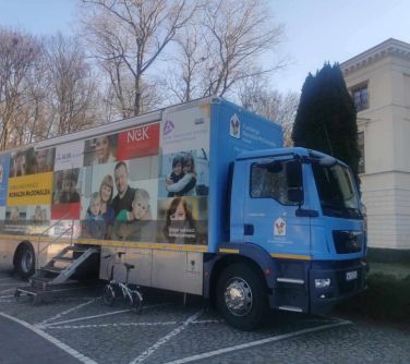 Na placu dzień. Oklejony ambulans. Schodki metalowe do wejścia. W tle drzewa