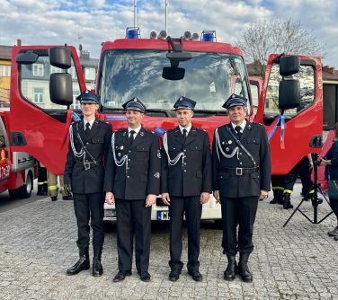 Na placu, dzień. Czterech strażaków w galowych strojach. Stoją przed nowym wozem strażackim