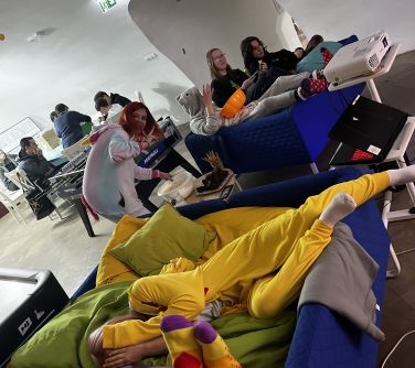 Pomieszczenie z białymi ścianami. Na kanapach siedzi młodzież. W tle na krzesłach