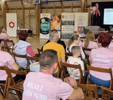 Oświetlona hala sportowa. na krzesłach sluchacze. Na przodzie scena z dwiema kanapami i stolikiem. Po lewej banery