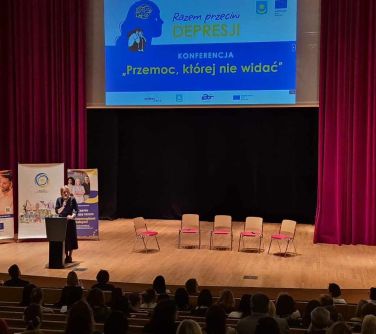 Oświetlona scena. na scenie pięć krzesel. Po lewej przy mównicy kobieta. W tle wisi duży ekran z tytułem razem przeciw...