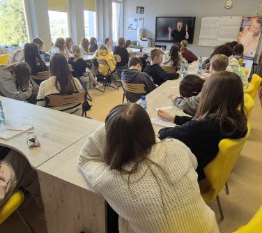 Jasna sala lekcyjna. Za oknem dzień. Przy stołach ustawionych w litere u siedzi młodzież. Na przodzie prowadzący zajęcia