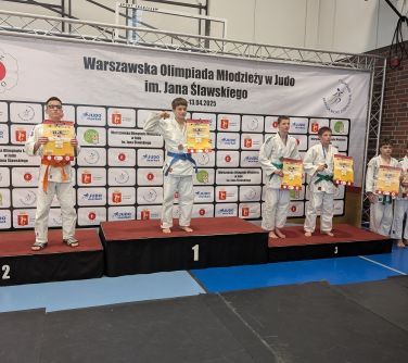 Oświetlona hala sportowa. Na podium czworo zawodników z dyplomami. W tle duży baner z logami
