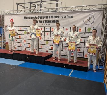 Oświetlona hala sportowa. Zawodnicy w białych kimonach na podium. W tle jasny baner z logami
