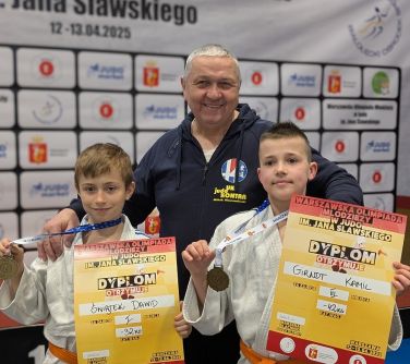 Uśmiechnięty mężczyzna. Przed nim dwóch młodych zawodników w bialych kimonach. Trzymaja w dłoniach medale i dyplomy