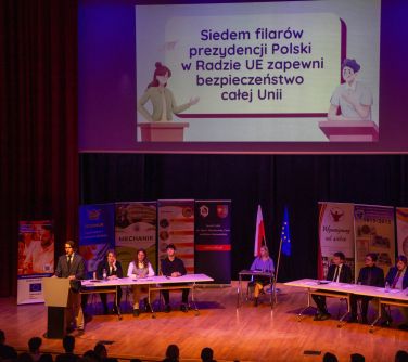 Aula, widok na scenę. Dwa długie stoły, po środku mały. Przy nich osoby. W tle stoją banery. Nad sceną wisi ekran z treścią