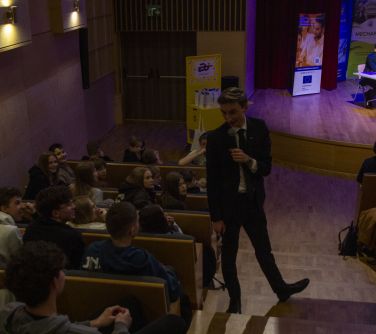 Aula, przyciemnione światło. Na schodach z mikrofonem mężczyzna. Zwraca sie do osób siedzących na fotelach