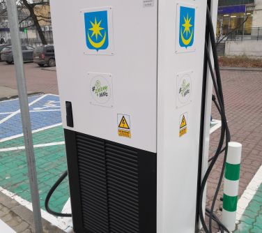 Na dworze, pochmurny dzień. Biało-zielona stacja ładowania pojazdów elektrycznych. Zdjęcie od tyłu. Na dole czarna kratka...