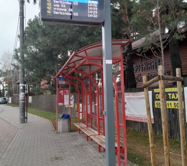 Na dworze, pochmurny dzień. Prz przystanku autobusowym elektroniczna tablica z rozkładem jazdy