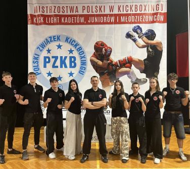 Oświetlona hala. Dziesięć osób w ciemnych strojach na tle baneru Mistrzostwa Polski w kickboxingu