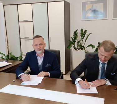 Jasne pomieszczenie. Przy stole na fotelach dwóch mężczyzn podpisuje umowę
