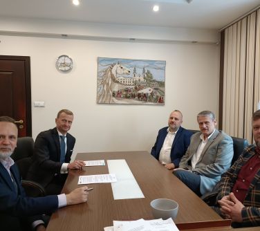 Jasne pomieszczenie. Pięciu mężczyzn siedzi przy stole. Na stole dwa egzemplarze umowy. Na przodzie na jasnej ścianie obraz...