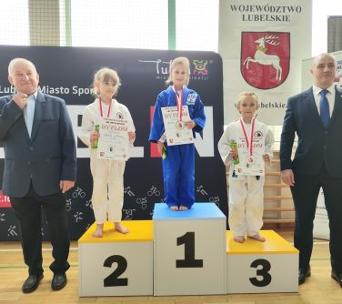 Jasna naturalnym światłem hala. Na parkiecie podium. Na nim trzy zawodniczki. Na pierwszym miejscu dziewczynka w niebieskim...