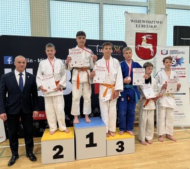 Jasna naturalnym światłem hala. Na parkiecie podium. Na nim zawodnicy. Po lewej stronie podium mężczyna w garniturze. Po...