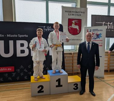 Jasna naturalnym światłem hala. Na parkiecie podium. Na nim dwóch zawodników. Po prawej stronie podium mężczyna w garniturze.