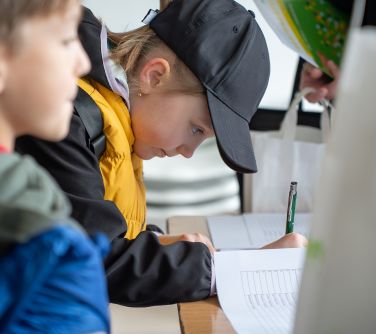 Pochylona dziewczynka w ciemno-żółtej bluzie i czapeczce basebolówce podpisuje listę. Obok dłoni butelka z wodą