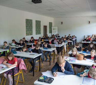 Jasna sala lekcyjna, światło naturalne z okien. Przy stolikach siedzi po dwoje dzieci. Piszą sprawdzian