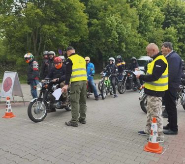 Na dworze, dzień. Dwa pachołki po dwoch stronach ulicy z kostki. Za nimi motocykliści na motorach