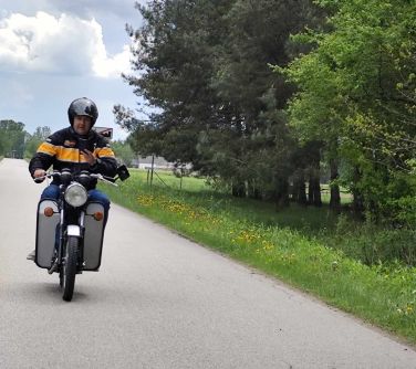 Na dworze, dzień. Motocyklista w kasku jedzie motoem po ulicy. Po prawej zielone drzewa i trawa