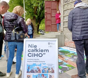 Na zewnątrz, dzień. Na przodzie stojąca ramka z plakatem ni ecalkiem ciho. Naokolo osoby. patrza na kolorowa matę rozlożoną...