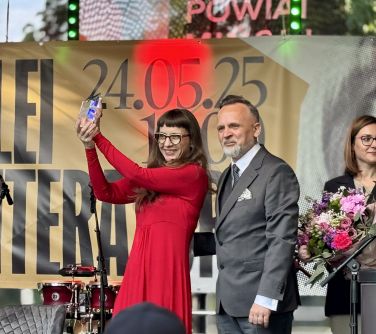 Na dworze, dzień, scena. Kobieta w czerwonej sukience podnosi statuetke do góry, obok mężczyzna