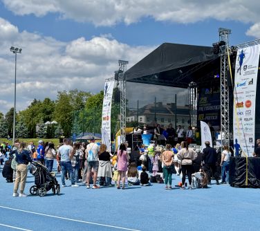 Na dworze, dzień, słońce. Na niebieskiej murawie osoby. Gromadzą się przed rozstawioną sceną