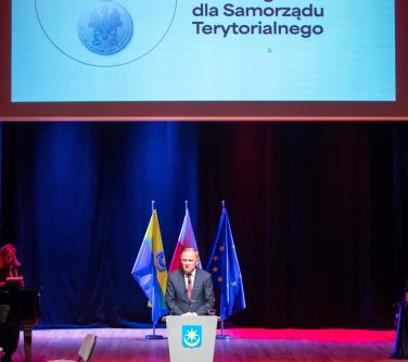 Oświetlona sala koncertowa. Widok na scenę. Na niej po środku mównica i przed nią kobieta. Wisi ekran z hasłem odznaka...