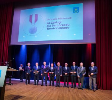 Oświetlona sala koncertowa. Widok na scenę. Na niej grupa odznaczonych mężczyzn w garniturach. Wisi ekran z hasłem odznaka...