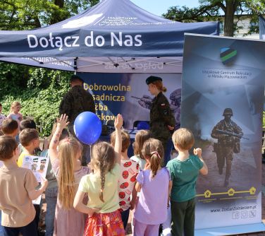 Na dworze, dzień, słońce. Grupa dzieci przed namiotem wojska. pod namiotem żołnierz i żołnierka. Po prawej baner z...