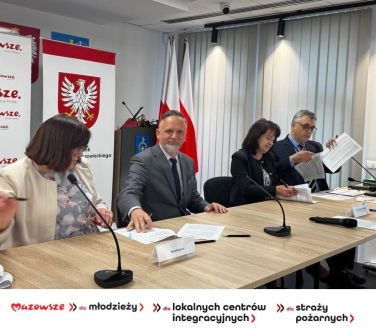 Oświetlone pomieszczenie. Przy stole cztery osoby podpisują dokumenty. W tle banery