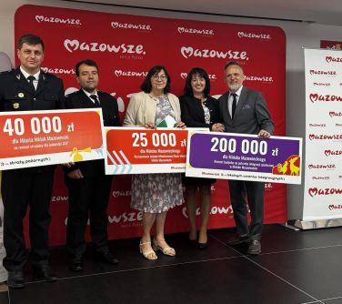 Oświetlone pomieszczenie. Pięć osób pozuje trzymając duże banery. Na nich kwoty. W tle czerwony duży baner z białymi...